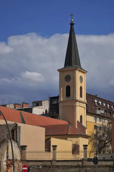 The church in Pančevo, a striking building with a tall, slender bell tower topped by a cross. The tower has a visible clock face. Adjacent to the church are various urban buildings with red and yellow facades, all under a dramatic cloudy sky.