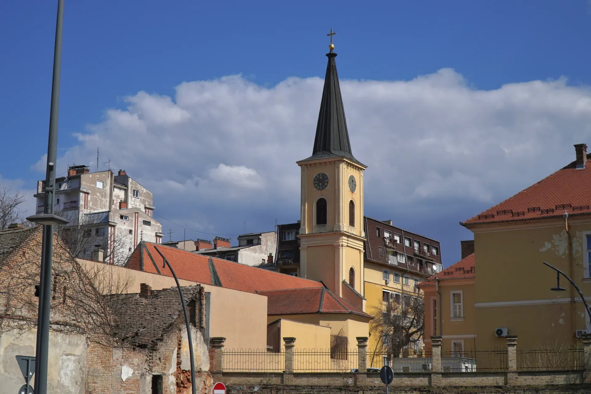 A yellow church tower with a black pointed roof and a clock is visible in Pančevo, Serbia. Modern buildings are in the background, and the sky is blue with white clouds.