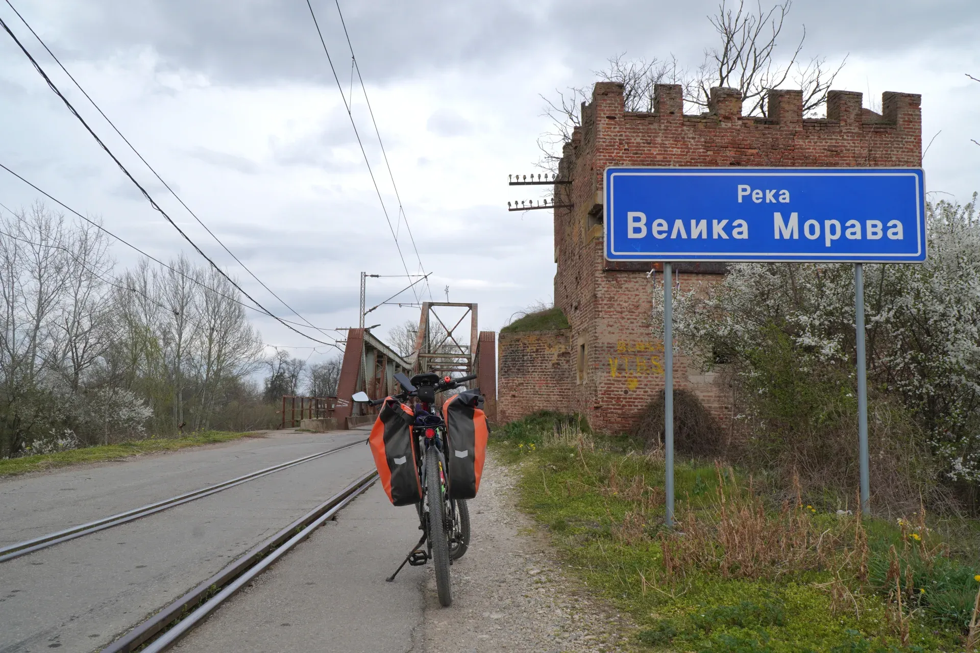 A bicycle loaded with orange saddlebags stands on a gravel path next to train tracks. In the background, the Ljubičevski Most (bridge) over the Great Morava is visible, along with an old brick tower and a blue sign that reads 'Velika Morava'. The scene is located near Požarevac, Serbia.