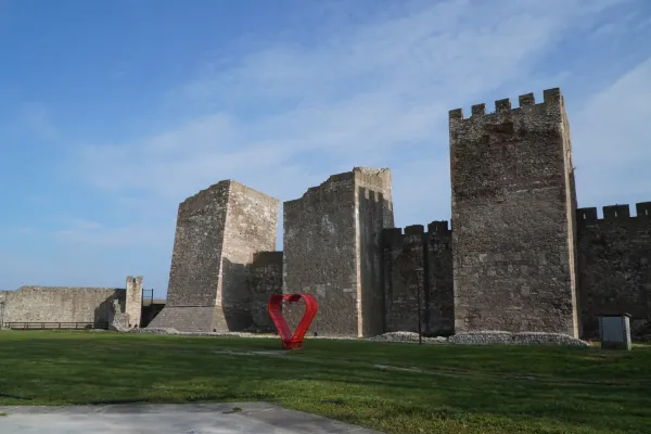 The Smederevo Fortress in Serbia with its tall stone walls and towers, next to it a bright red heart sculpture on a green lawn.