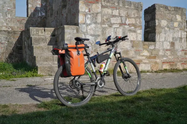 A bicycle loaded with orange panniers stands on a paved path in front of an old stone wall of Smederevo Fortress, Serbia.