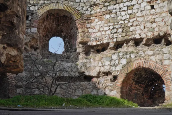 A dilapidated stone wall with several arches and openings through which the blue sky is visible, as well as dry branches and green vegetation on the ground.