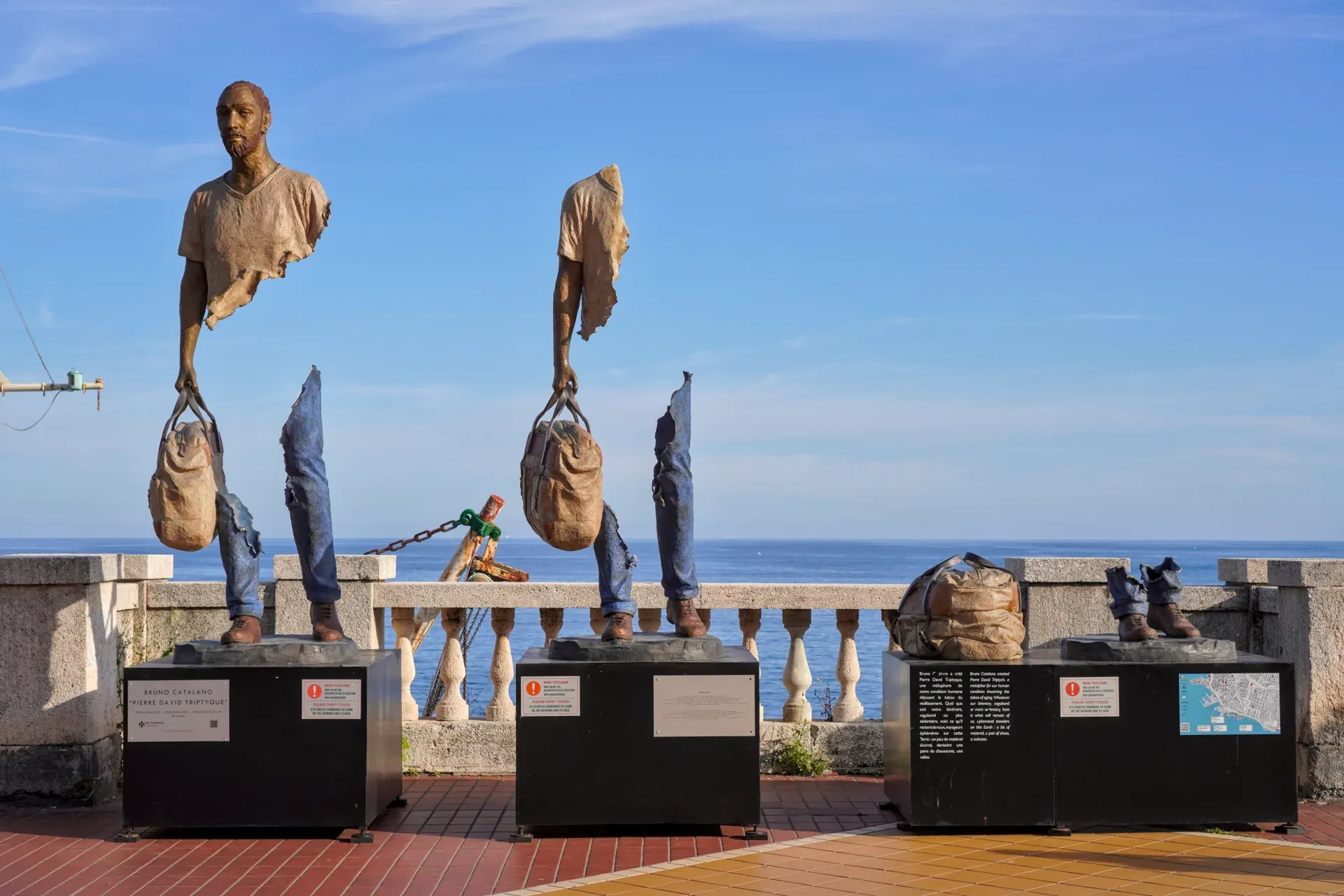 Sculptures fragmentées en bronze de torses et jambes humaines portant des sacs, exposées sur des socles le long de la promenade de Gênes, avec la mer bleue et le ciel en arrière-plan.