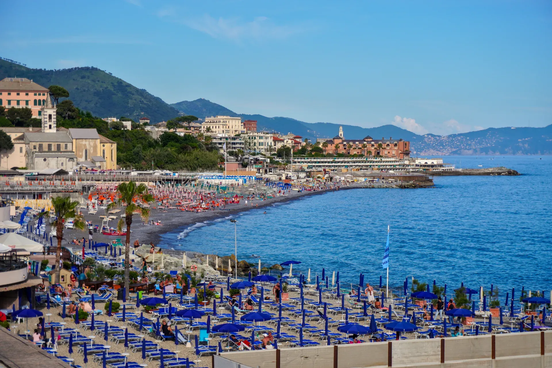 Uzbunjena plaža u Đenovi, Italija, sa brojnim plavim suncobranima i ležaljkama, zgradama i zelenim brdima u pozadini, i plavim morem sa desne strane.