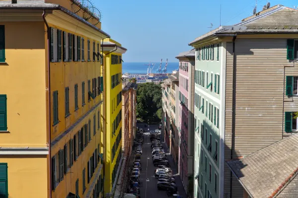 Eine bunte italienische Gasse mit pastellfarbenen Gebäuden, Autos und Blick auf das Meer und Hafenkräne.