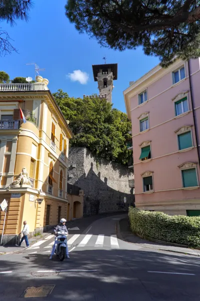 Un'imponente torre italiana si erge sopra alberi e edifici pastello a Genova, con uno scooter in una strada in primo piano.