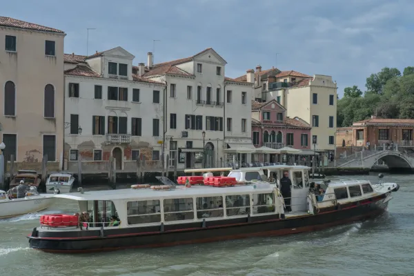 Vaporetto plovi kanalom u Veneciji, okružen istorijskim zgradama i pešačkim mostom u pozadini.