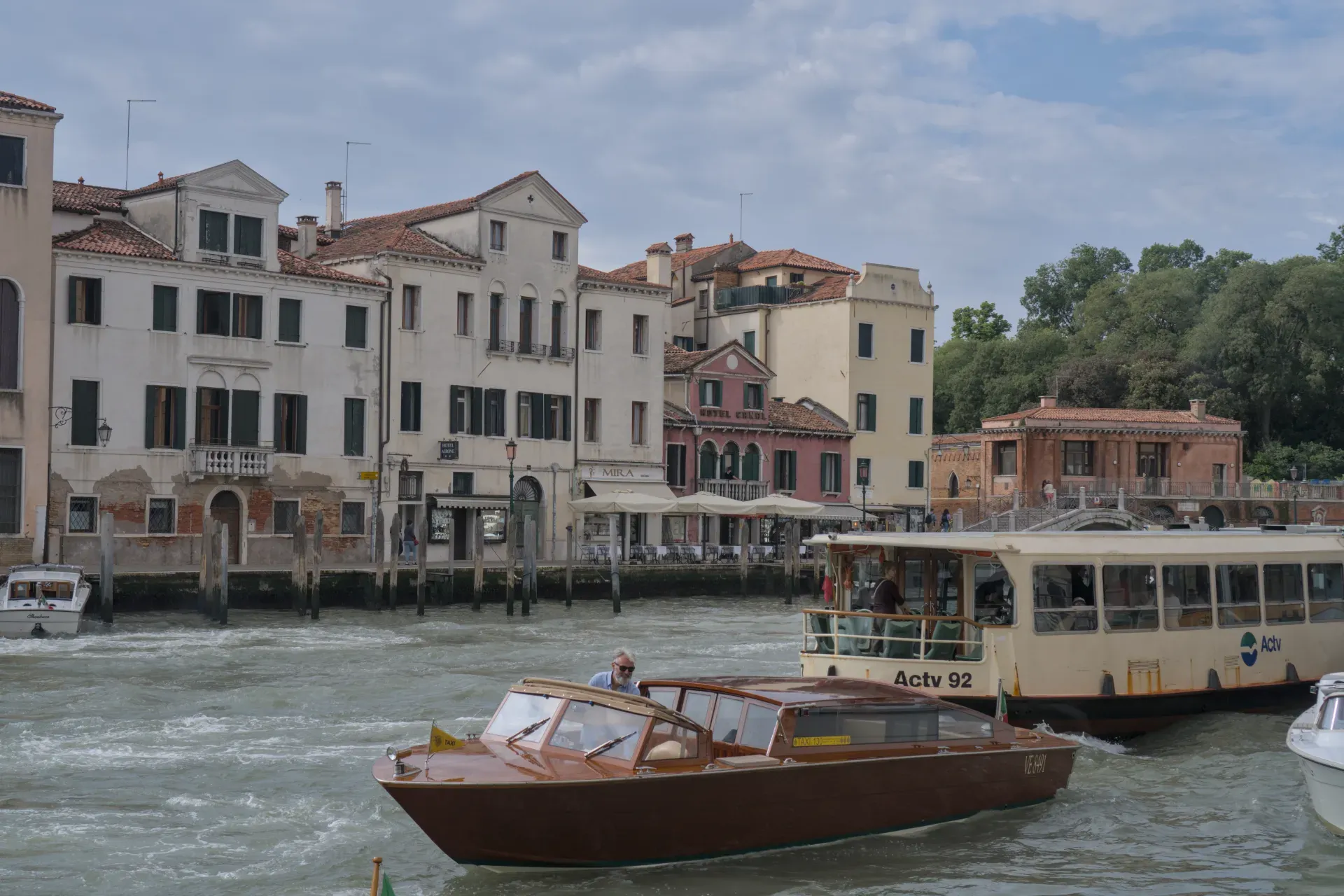 A Venetian canal with a brown taxi boat front view and a larger passenger ship, surrounded by classic buildings under a cloudy sky.
