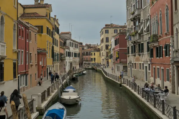 Eine enge Gasse in Venedig mit bunten Häusern, einem Kanal und Booten, mit Menschen, die auf einem Gehweg spazieren gehen.