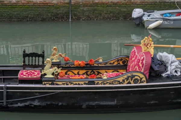 Eine prunkvolle venezianische Gondel mit roten und goldenen Verzierungen, treibend auf ruhigem, grünem Wasser vor einer Ziegelmauer.