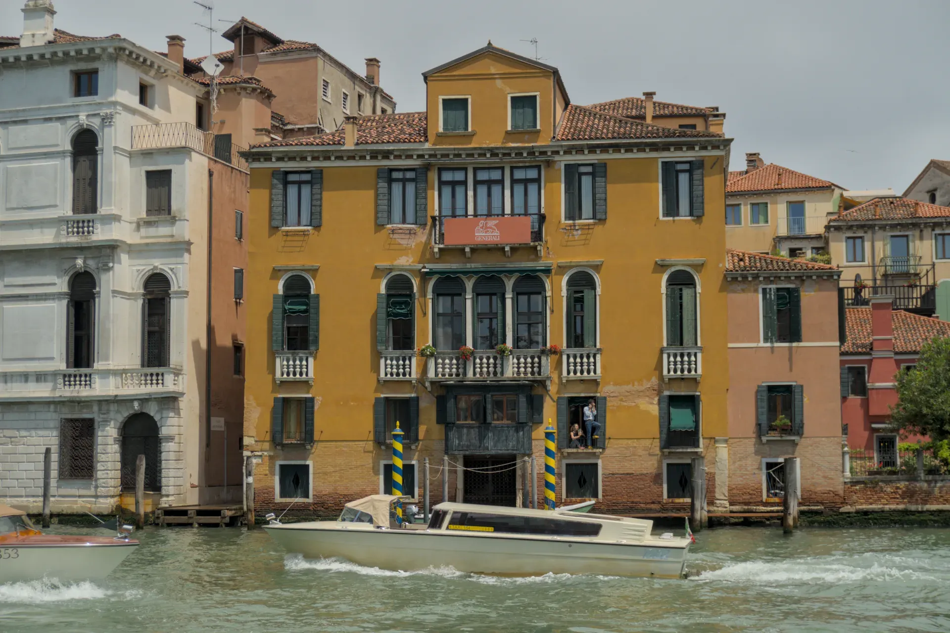 Ein Boot fährt auf einem Kanal in Venedig, vorbei an einem venezianischen Palazzo mit einer beigen Fassade und grünen Fensterläden.
