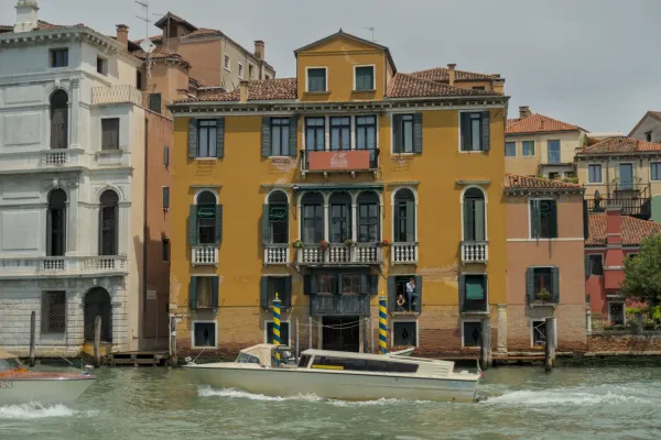 Ein Boot fährt auf einem Kanal in Venedig, vorbei an einem venezianischen Palazzo mit einer beigen Fassade und grünen Fensterläden.