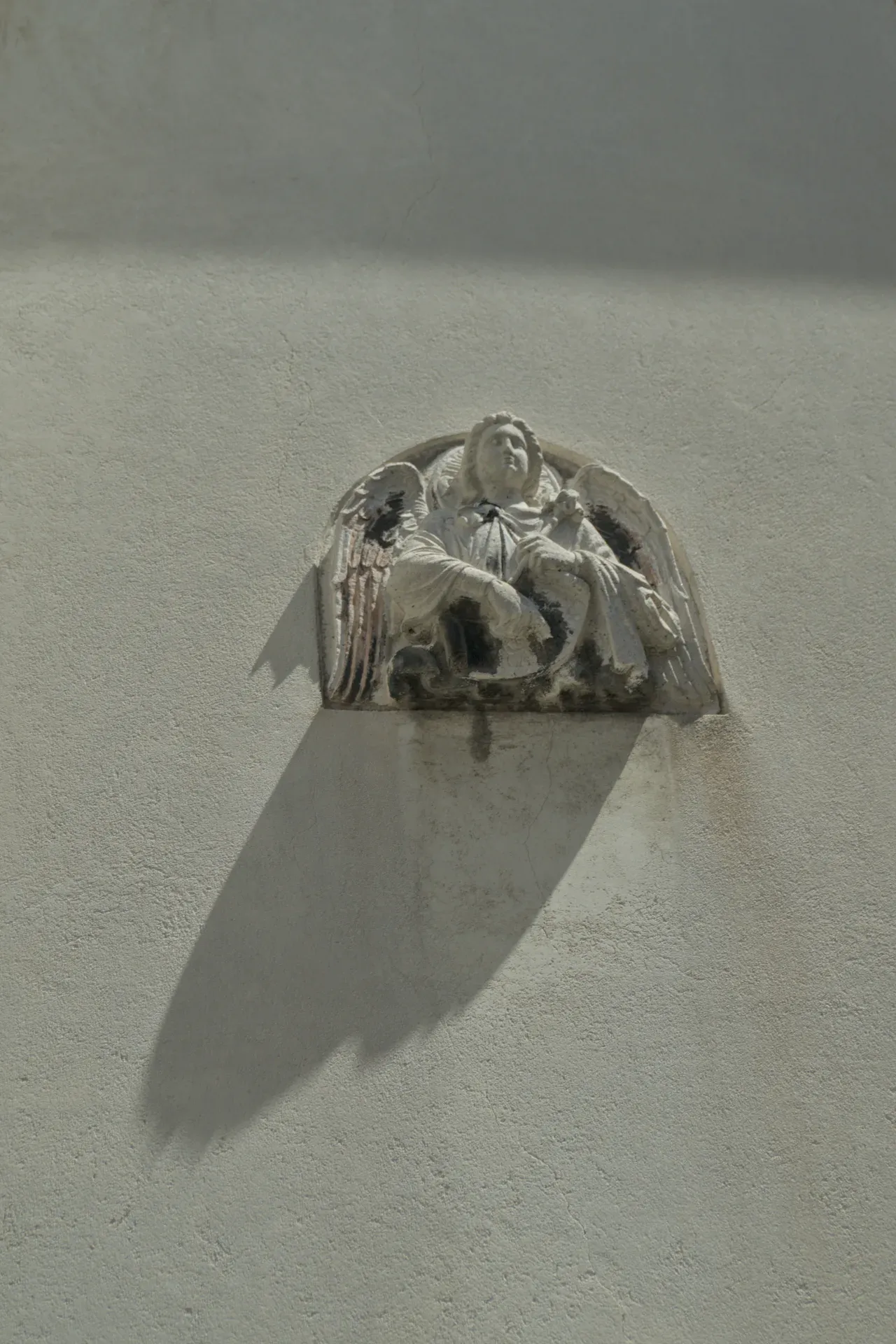Sculpture of an angel with wings on a textured, light wall with a dramatic shadow.