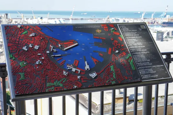 Una dettagliata mappa in modello 3D di Genova, Italia, che mostra l'architettura della città e l'area del porto, con il Mar Ligure e le gru portuali sullo sfondo.