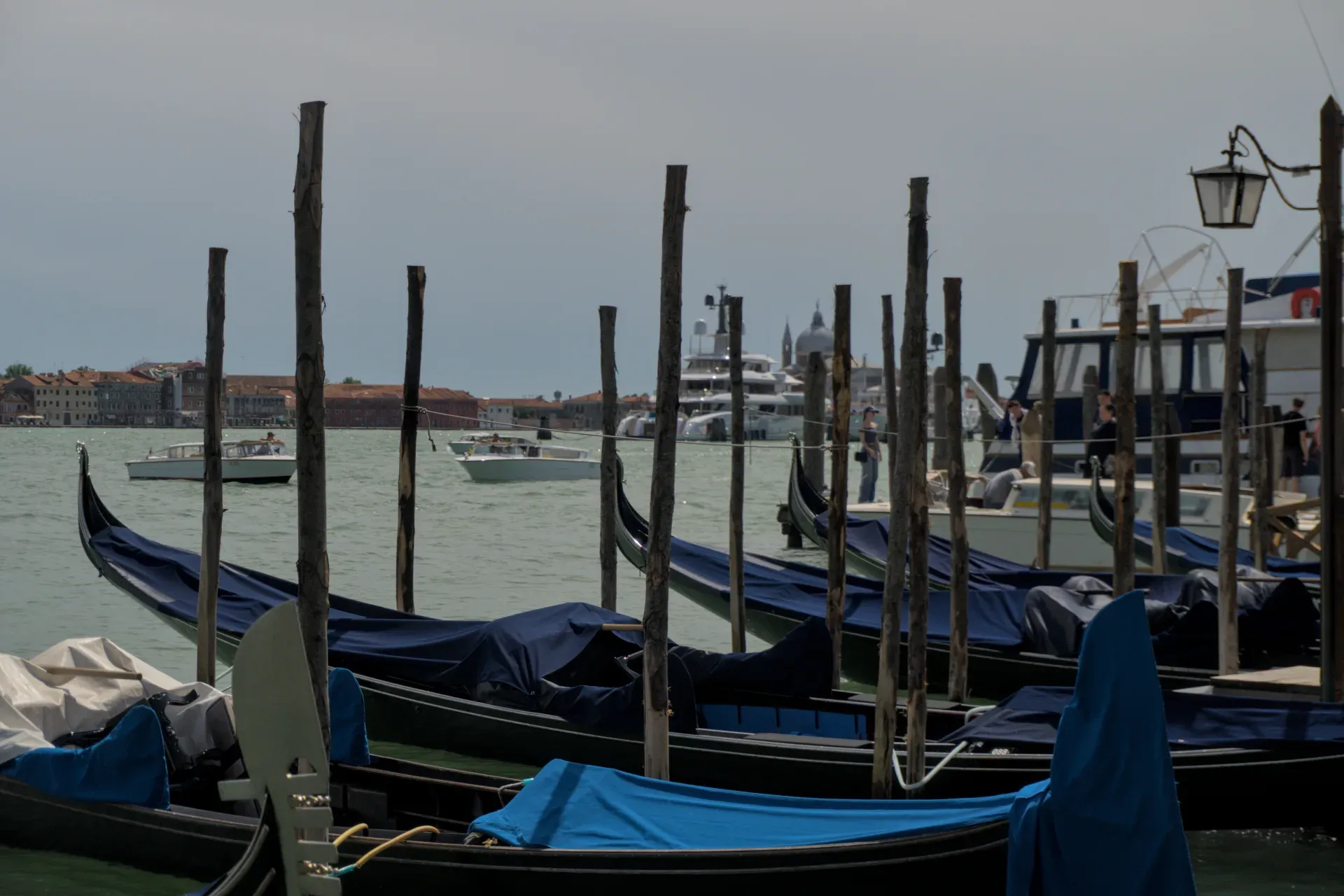Eine Reihe von venezianischen Gondeln, mit blauen Abdeckungen, vor der Kulisse der Stadt und des Wassers, mit anderen Booten im Hintergrund.