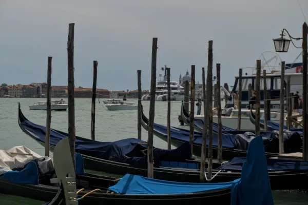 Eine Reihe von venezianischen Gondeln, mit blauen Abdeckungen, vor der Kulisse der Stadt und des Wassers, mit anderen Booten im Hintergrund.