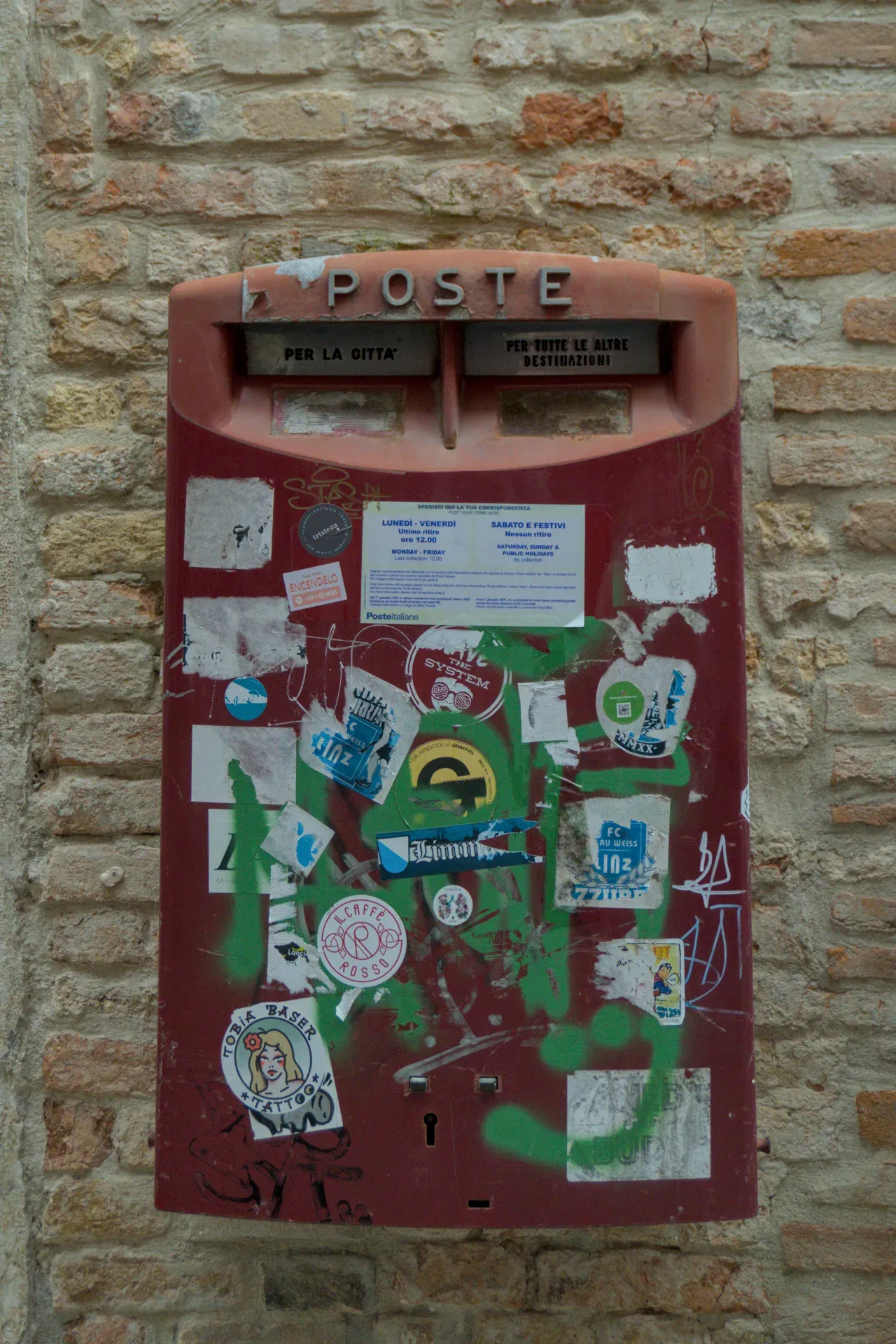 An old red postbox decorated with stickers and graffiti against a brick wall in Venice.