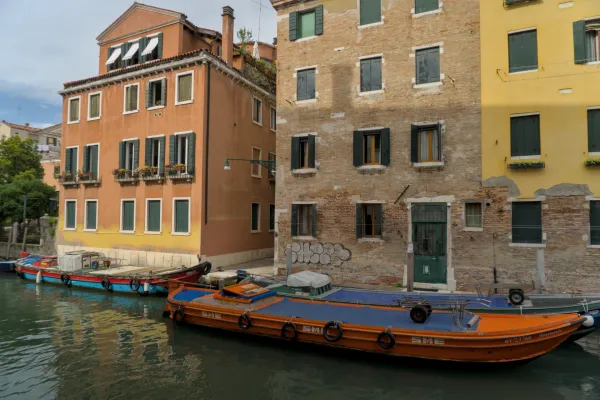 Pogled na venecijanske kanale sa čamcima ispred istorijskih narandžastih i žutih zgrada sa zelenim kapcima.