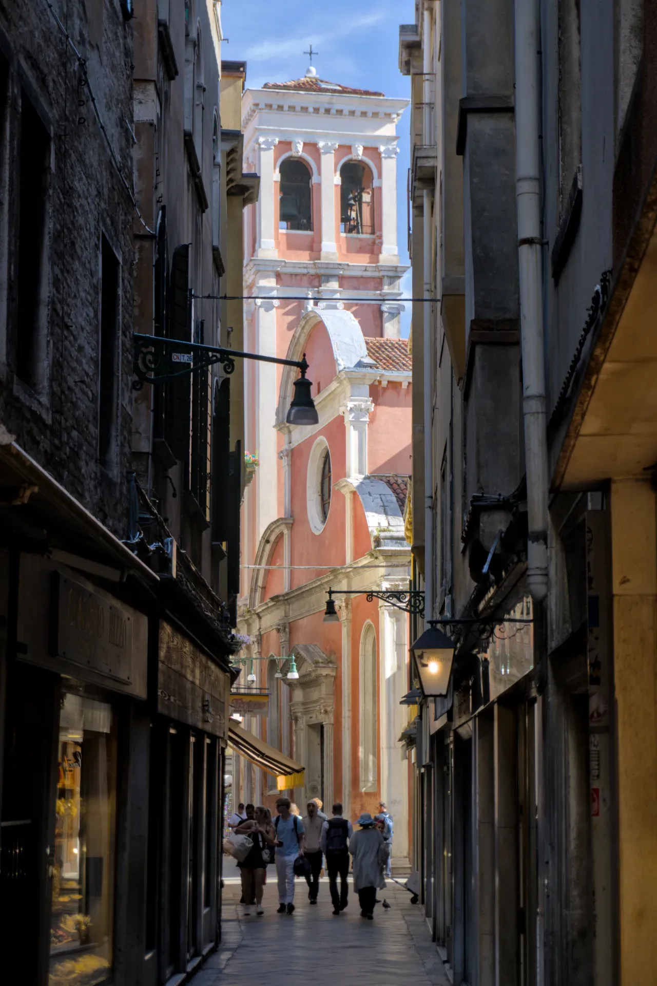 Fotografia di uno stretto vicolo a Venezia che conduce a un campanile rosa con campane e archi, con persone che camminano.
