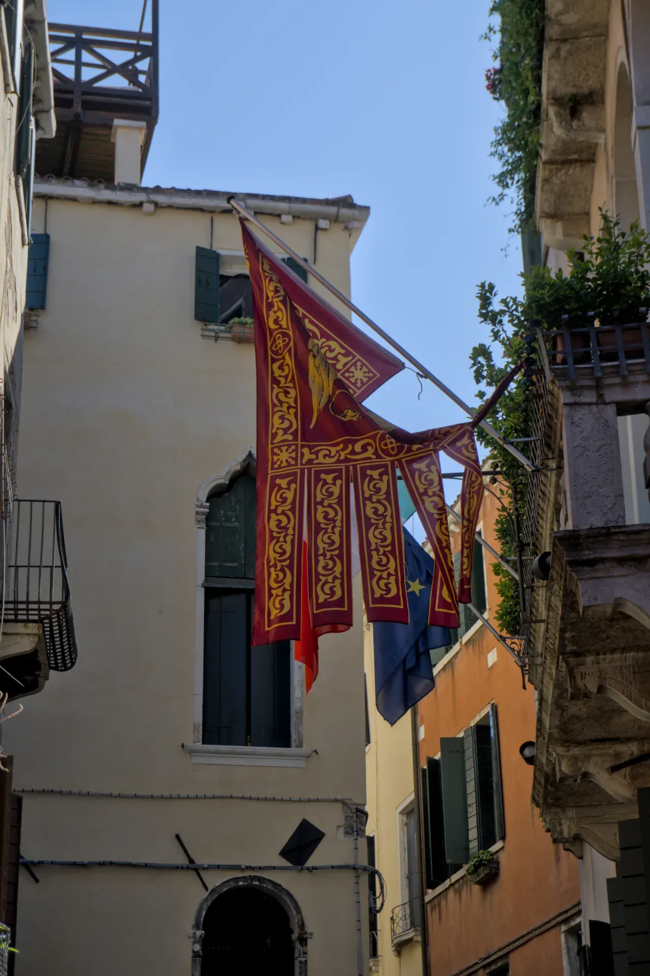 Venetian flags with golden embellishments wave in the sunlight in front of beige and orange building facades in an alley.