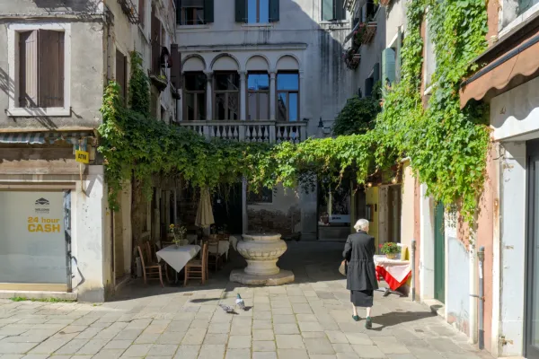 Una donna cammina attraverso un vicolo veneziano verde e coperto d'edera, con tavoli all'aperto e un pozzo antico.