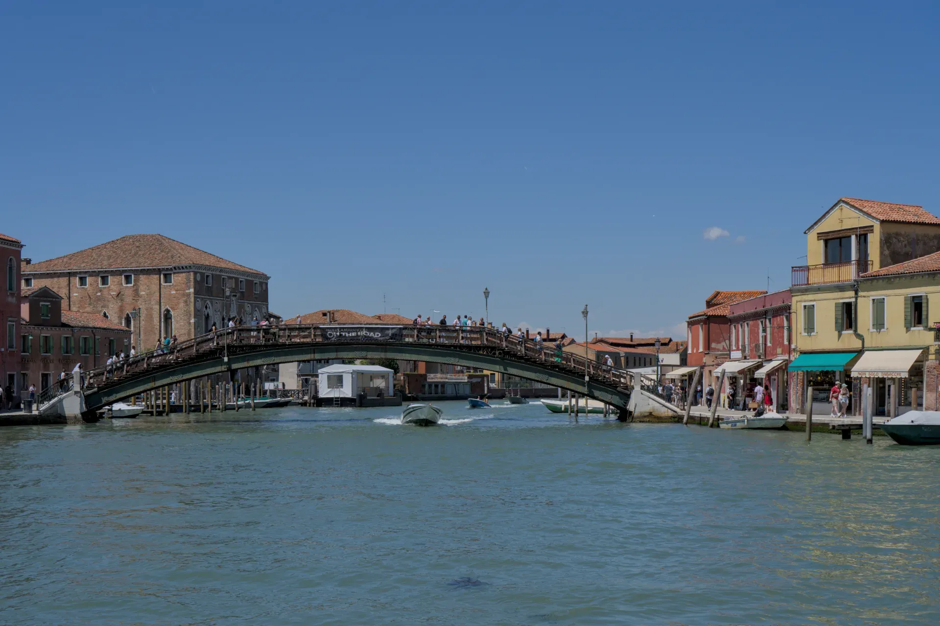 Eine hölzerne Fußgängerbrücke auf der Insel Murano in Venedig, überquert von Menschen, mit Booten auf dem Wasser und historischen Gebäuden.