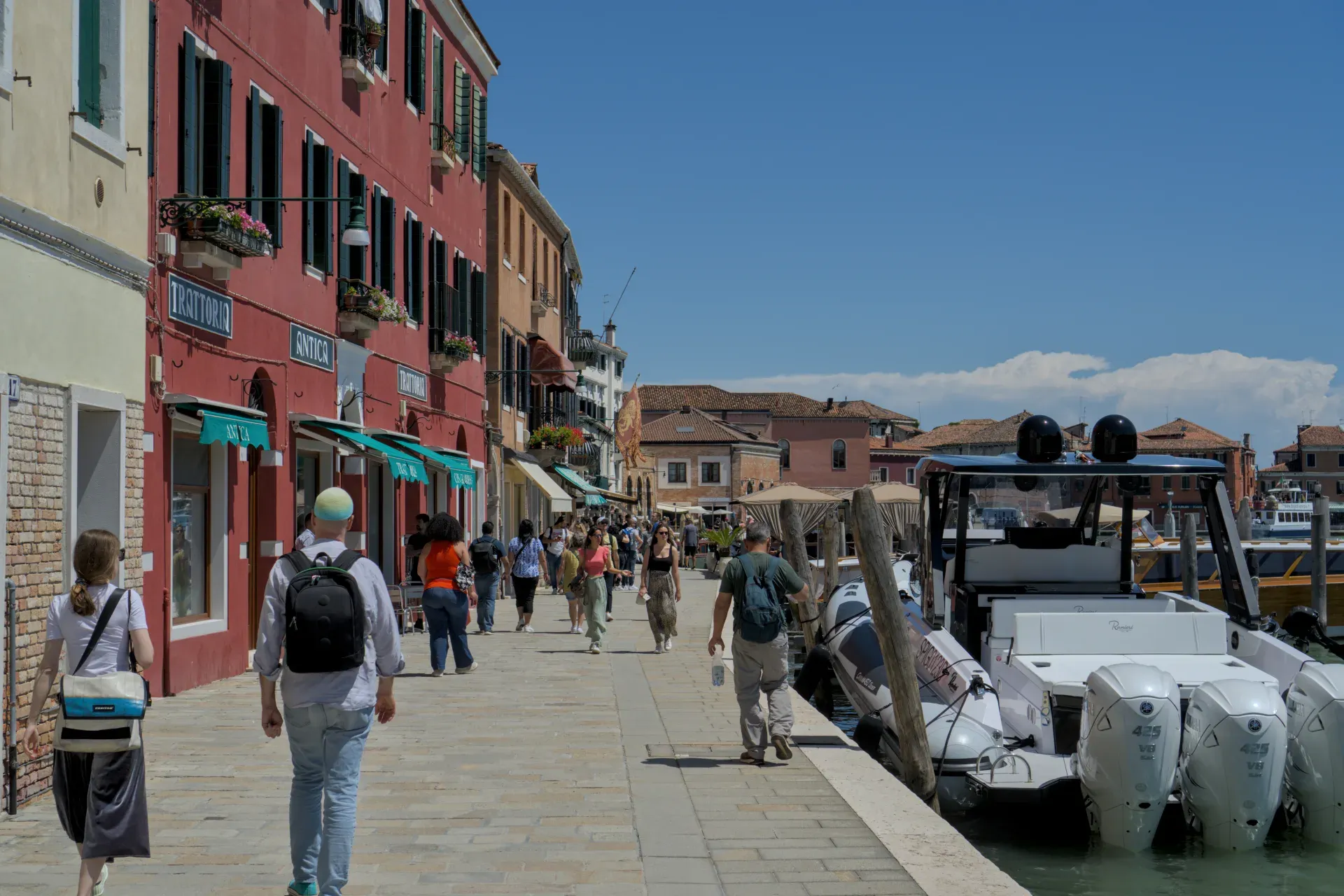Gente che passeggia lungo la soleggiata riva dell'isola di Murano, Italia, con case colorate e barche sull'acqua.