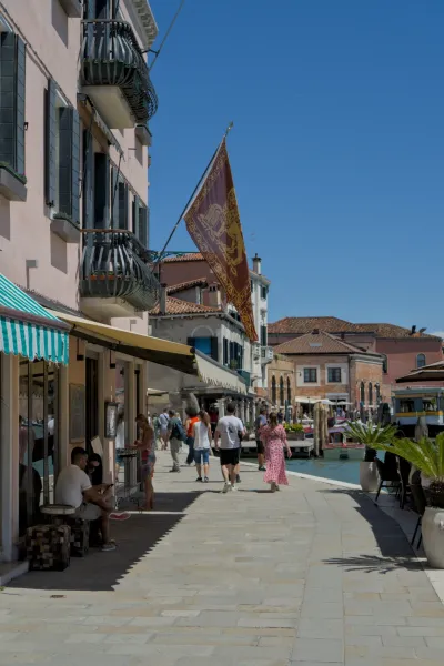 Persone passeggiano lungo un canale sull'isola di Murano, Venezia, sotto una bandiera veneziana sventolante.