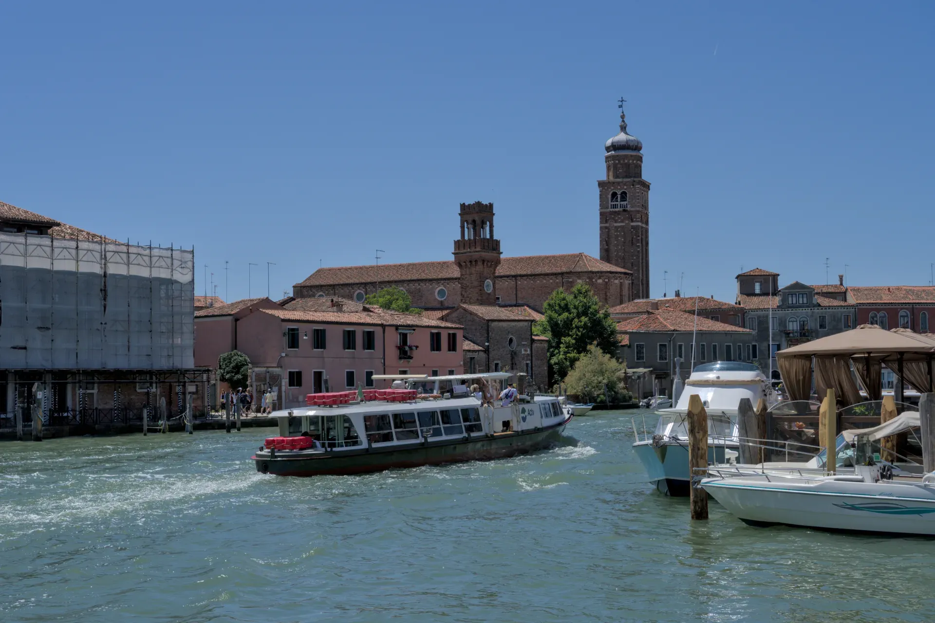 Traffico di barche sul canale dell'isola di Murano, Venezia, con edifici storici di chiese sullo sfondo alla luce del sole.