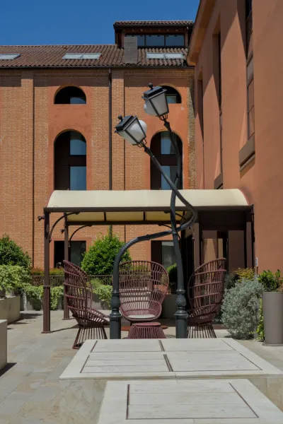 Eine sonnige Terrasse auf Murano mit Korbstühlen und einem stilvollen Lampenposten vor Backstein- und Terrakotta-Gebäuden.
