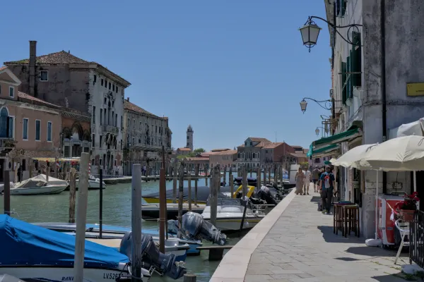 Pogled na kanal u Veneciji sa čamcima usidrenim uz pristanište i tradicionalnim kućama u pozadini, sa Muranom u daljini
