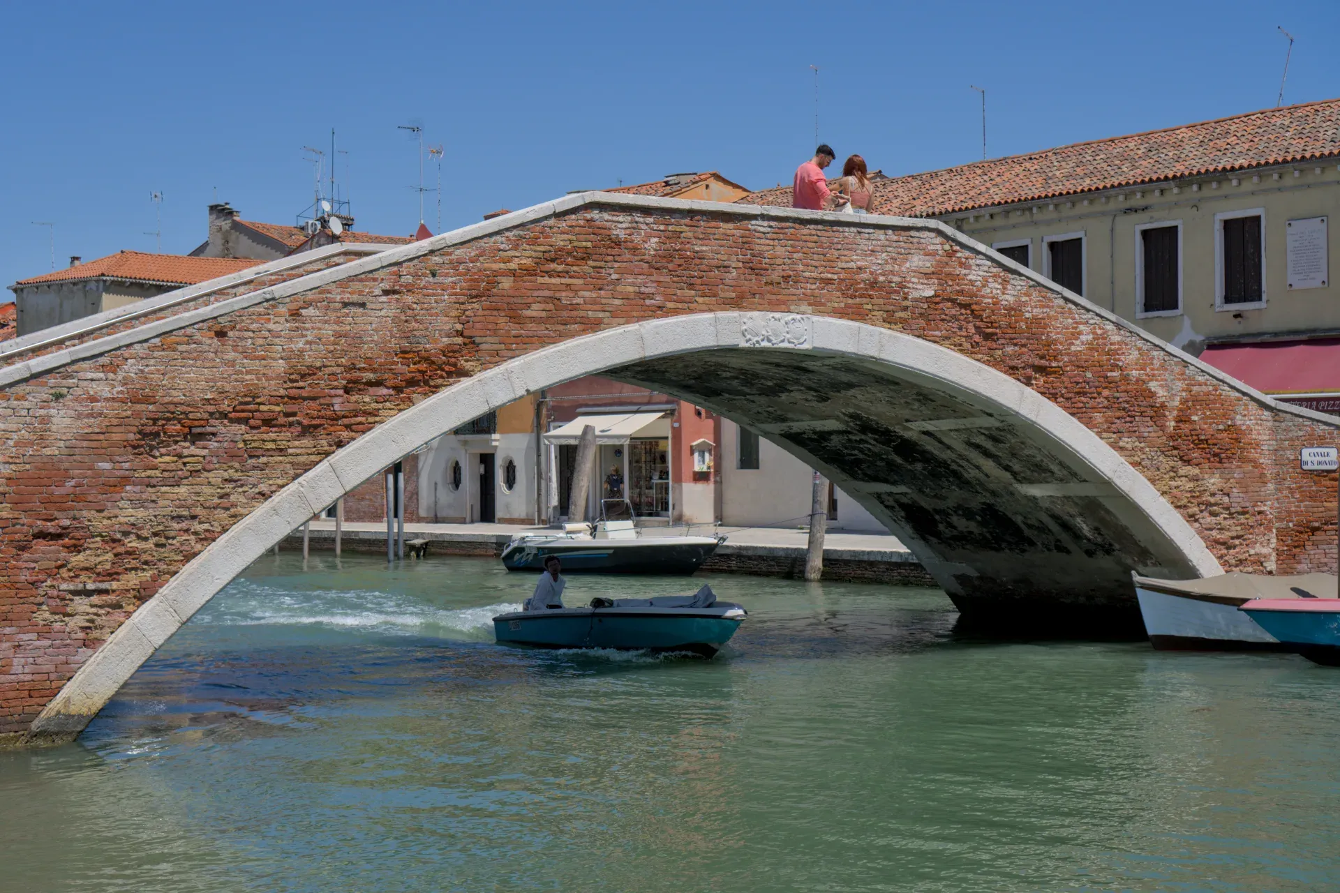 Eine steinerne Brücke mit Ziegelakzenten überspannt einen Kanal auf der Insel Murano, Italien, mit Booten unter der Brücke und einem Paar auf der Brüstung.