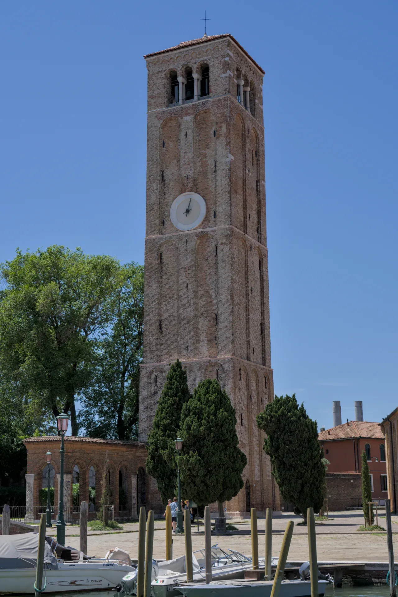 The brick bell tower of Murano island under a blue sky, with trees and water in front.
