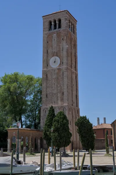 Der Backsteinglockenturm der Insel Murano unter blauem Himmel, mit Bäumen und Wasser davor.