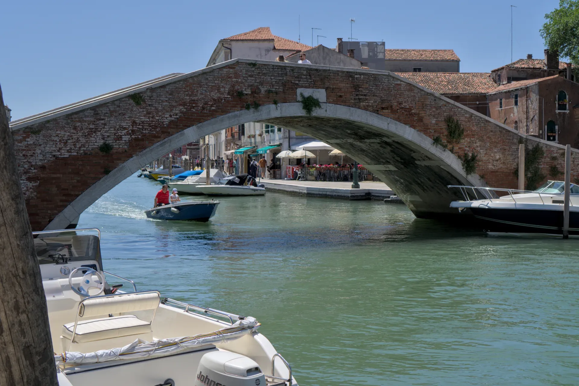 Historische Backsteinbrücke auf der Insel Murano, Venedig, überspannt einen Kanal mit kleinen Booten und Cafés.
