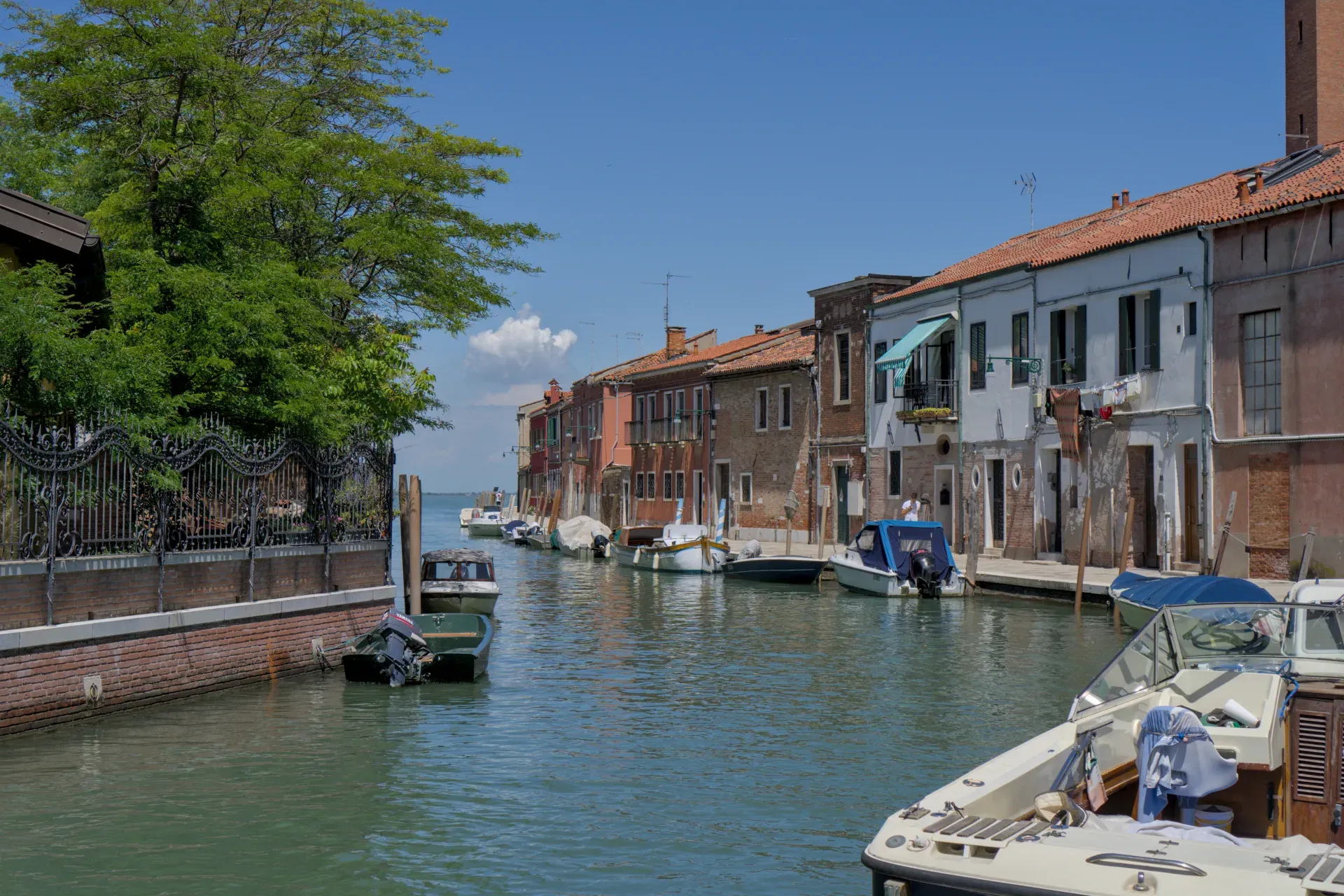 Kanal na Muranu, Italija, obrubljen šarenim kućama i čamcima, pod vedrim plavim nebom.