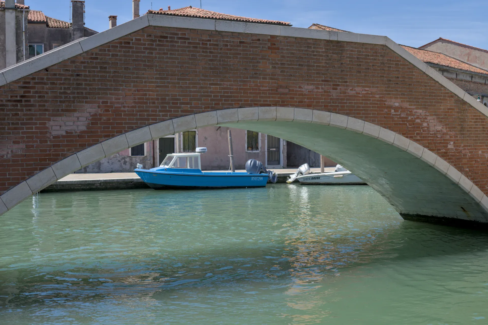 Un ponte in mattoni rossi con decorazioni in pietra sovrasta un canale, con una barca blu adagiata su di esso, vicino all'isola di Murano.