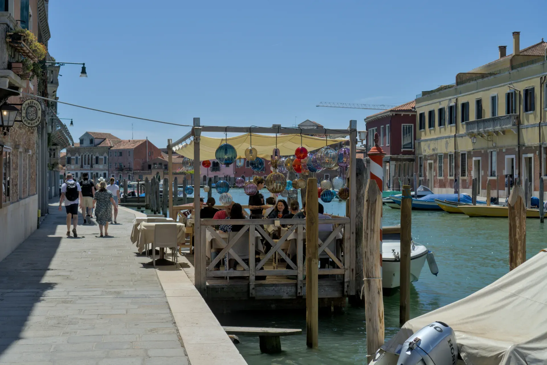 Ein Kanal in Murano mit einem Restaurant und bunten Glaslichtern, im Hintergrund traditionelle Gebäude und Boote.
