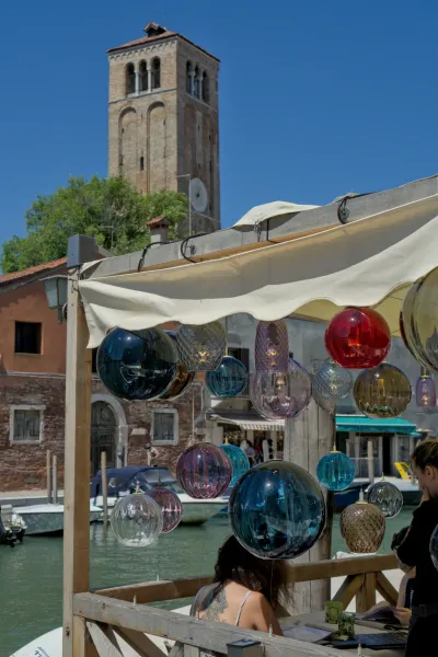 Handgefertigte Murano-Glasleuchten hängen über einem Kanal in Venedig mit einem Turm im Hintergrund.
