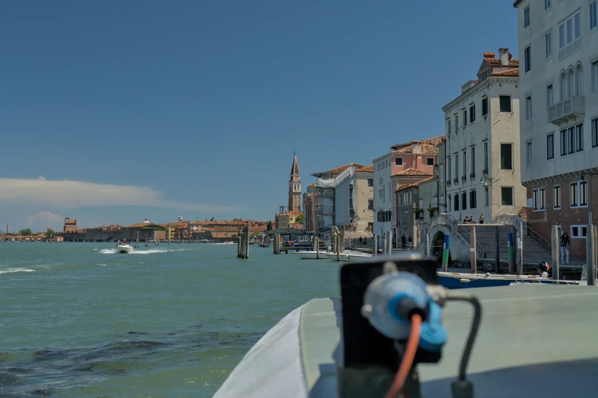 Ein sonniger Tag in Venedig, Blick auf das Kanalufer mit traditionellen Gebäuden, die sich im türkisfarbenen Wasser spiegeln, und einem vorbeifahrenden Boot.