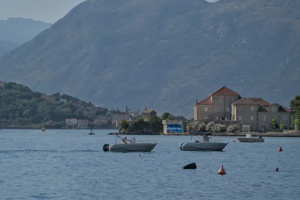Two boats on the calm blue waters of the Bay of Kotor, Montenegro, with a village and mountains in the background.