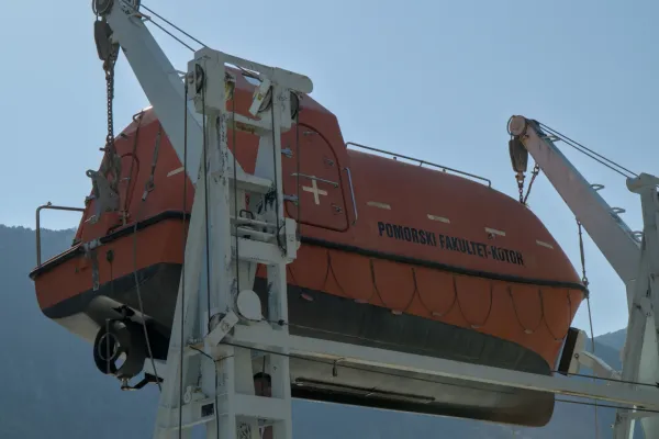 An orange lifeboat hangs from a crane over the water, with a blue sky in the background.