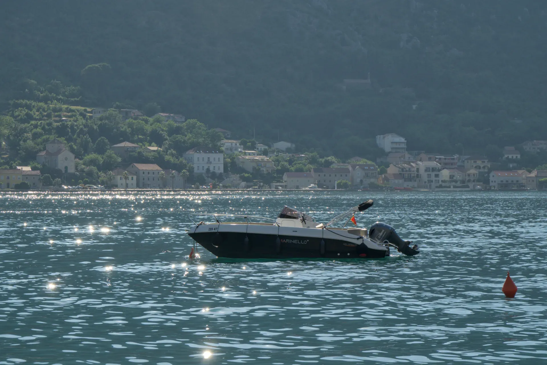 Una barca bianca e nera galleggia sulle acque soleggiate della Baia di Kotor, con una città costiera e colline sullo sfondo.