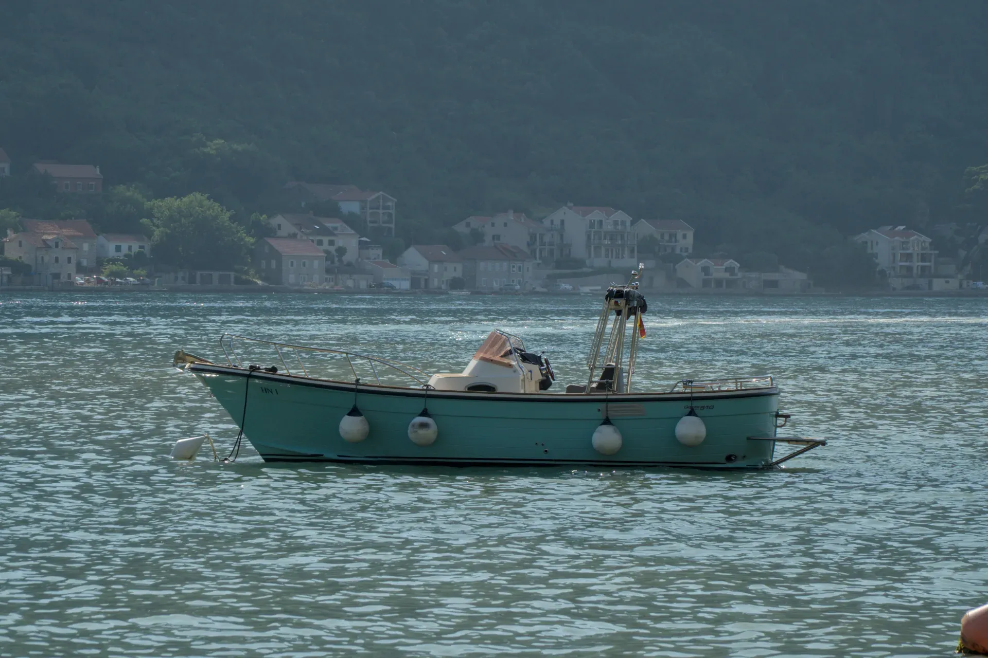 Una barca turchese con parabordi bianchi sul fianco galleggia su acqua calma di fronte a una città costiera a Kotor, Montenegro.