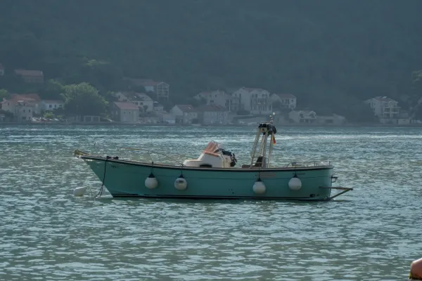 A turquoise boat with white fenders on its side floats on calm water in front of a coastal town in Kotor, Montenegro.