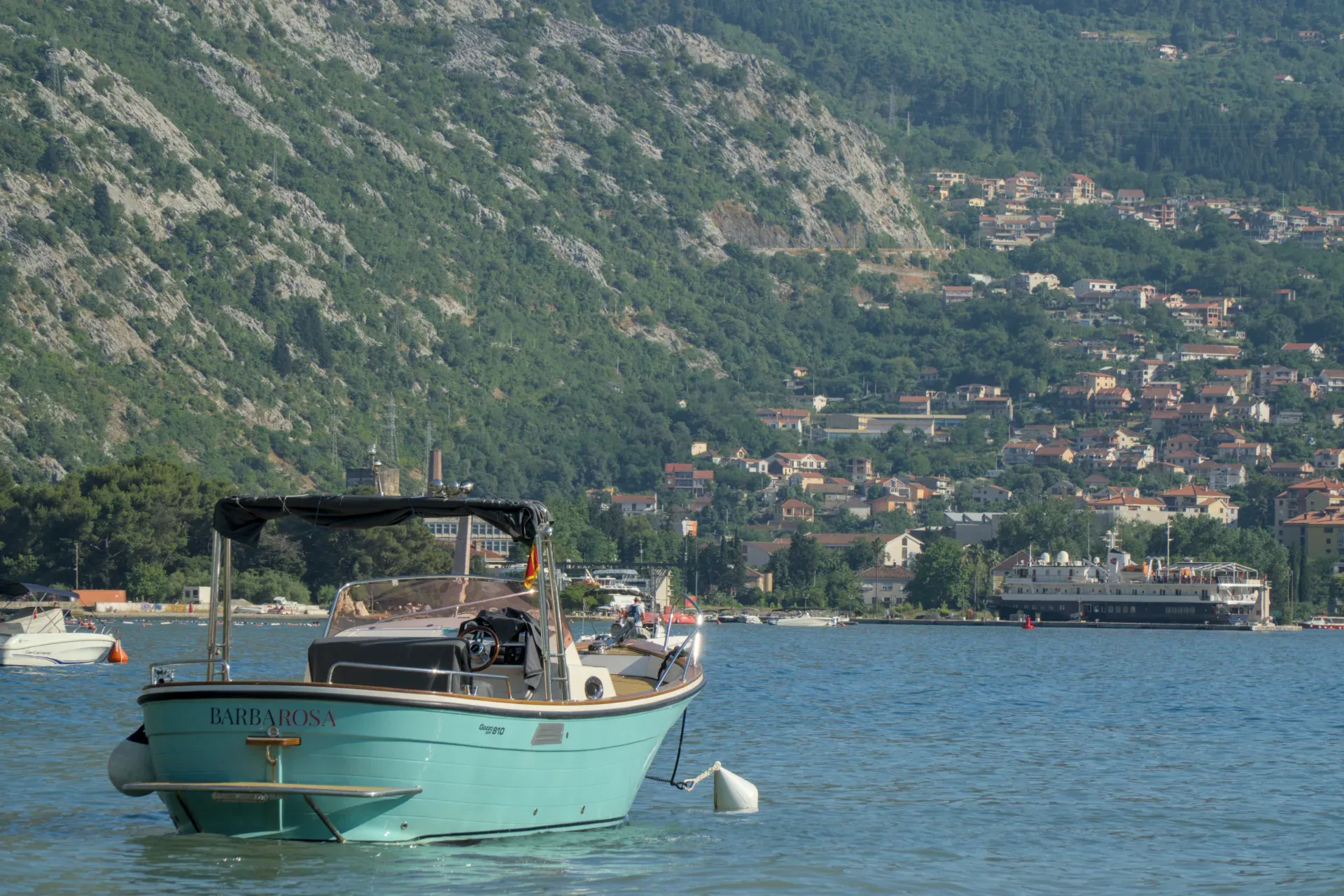 Una barca turchese riposa al largo della costa della Baia di Kotor, Montenegro, con colline verdi sullo sfondo.