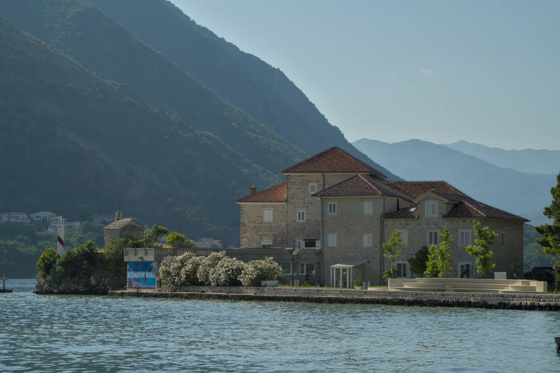 Edificio storico sulla Baia di Kotor con montagne sullo sfondo e una struttura simile a un faro.
