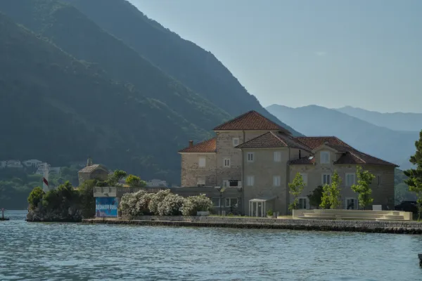 Historic building on the Bay of Kotor with mountains in the background and a lighthouse-like structure.