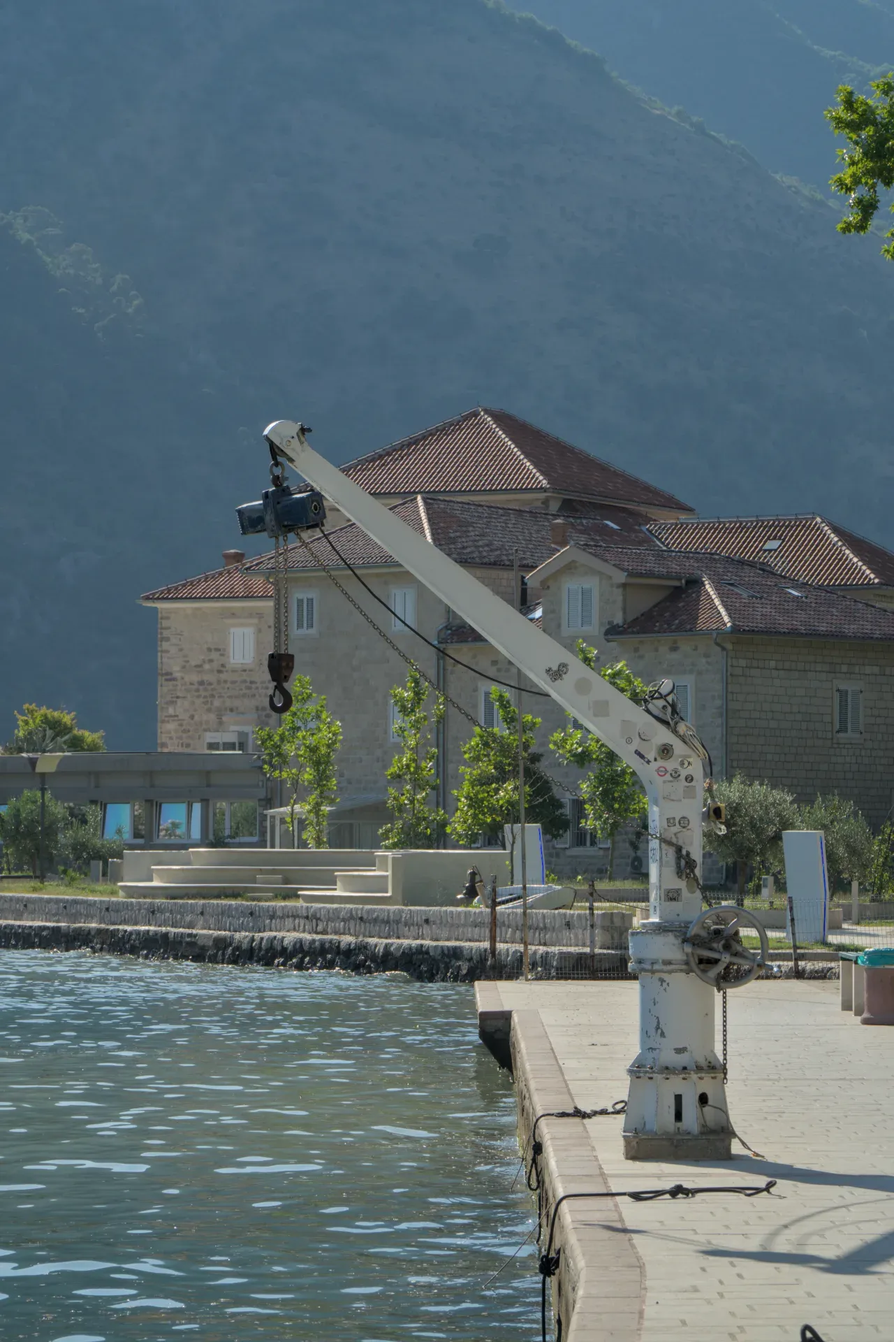 Ein industrieller Kran am Ufer der Bucht von Kotor, mit einem Haus und einem Berg im Hintergrund.