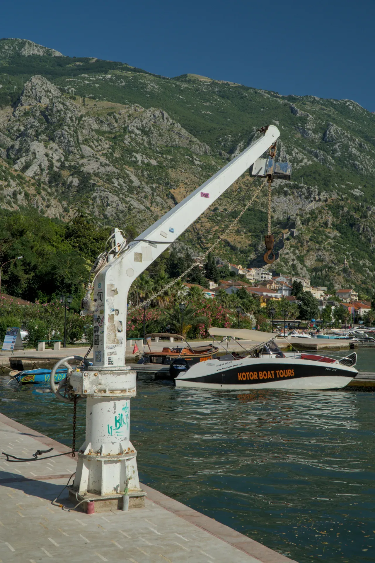 Una grúa portuaria blanca se alza en primer plano, con barcos y la ciudad de Kotor al fondo, contra un paisaje montañoso.